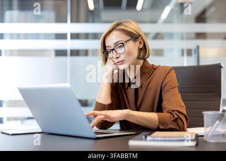 Young businesswoman wearing glasses and a brown shirt working with concentration on a laptop in a modern office, symbolizing business, technology, and professional focus Stock Photo