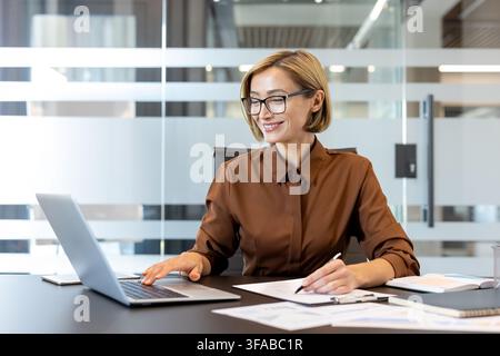 Businesswoman with short blonde hair and glasses smiling, typing on a laptop and making notes on a document while sitting at her desk in a modern corporate office environment Stock Photo