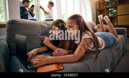 Two school-age girls browsing laptop content together while lying on sofa Stock Photo
