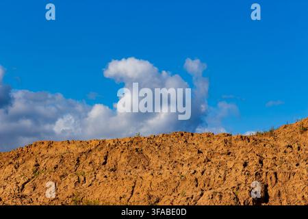 Large piles of excavated brown soil and raw earth mounds set against a vibrant blue sky with fluffy white clouds, showcasing raw land development and Stock Photo