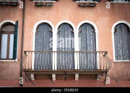 04 October 2022: View of a traditional Venetian building facade featuring arched windows with weathered blue wooden shutters and a decorative wrought- Stock Photo