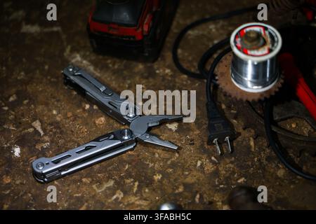 A close up of multitool, electrical plug, power tool, and metal component on worn workshop surface, creating rugged repair setting with practical indu Stock Photo