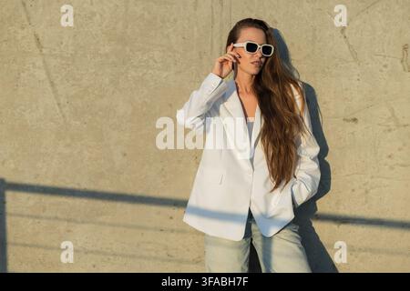 Stylish woman in white blazer and sunglasses touching her frames while leaning against a concrete wall. Horizontal medium shot in warm sunset light wi Stock Photo
