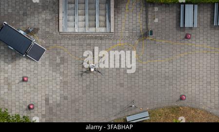 Aerial view of a drone connected by a yellow cable to a trailer on a paved plaza with benches and traffic cones in Amersfoort, Utrecht, Netherlands. Stock Photo