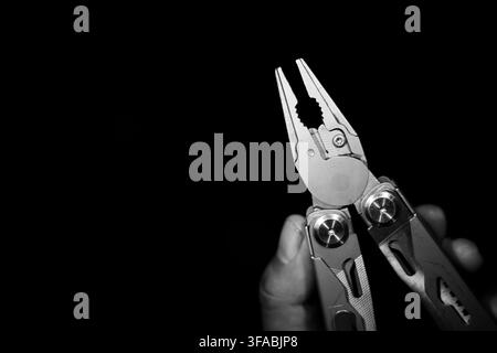 A pliers tool held by hand against black background, showing polished metal multitool with open jaws and dramatic high contrast for repair work Stock Photo