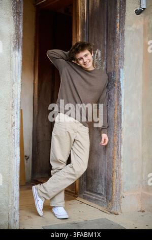 Casual youthful portrait. Smiling man with relaxed stance. Confident young man leaning against door in cozy attire. Urban styled young man casually posed with warm smile and relaxed demeanor Stock Photo