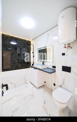 Bathroom interior featuring wall mounted sink and bathtub with shower system. White marble surfaces with black accent wall and water heater installed, concept of modern housing and utility efficiency Stock Photo