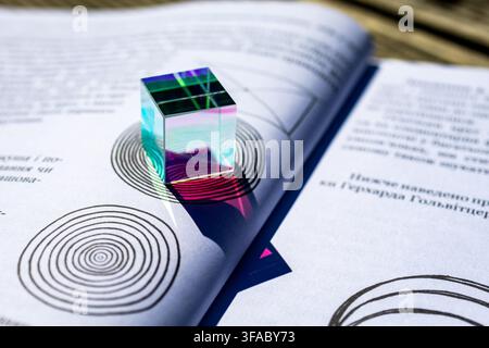 Colorful glass cube prism on an open book with concentric circle graphics. Sunlight produces vivid reflections and gradients. Concept for optics educa Stock Photo