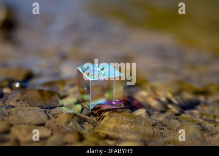 Shiny prism cube sits in clear moving water over stones. Surfaces glow with spectral color while the stream blurs into bokeh suggesting exploration mi Stock Photo