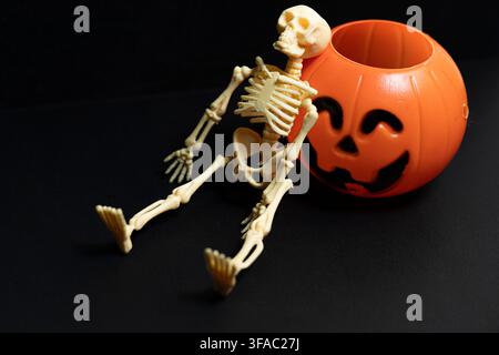 Small plastic skeleton sitting and leaning against an orange halloween pumpkin bucket on a dark background Stock Photo