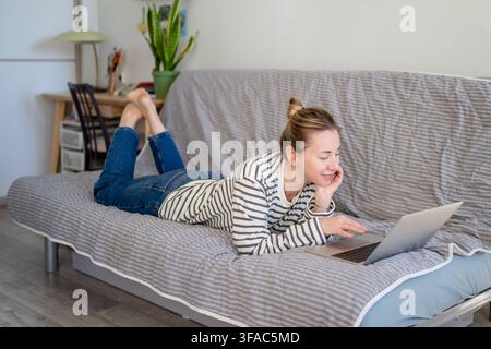 Cheerful curious woman browsing online on laptop, watching videos while lying on sofa at home. Stock Photo