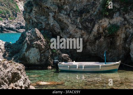 Traditional white wooden fishing boat moored in the clear turquoise waters of a rocky Greek coastal cove beneath limestone cliffs Stock Photo