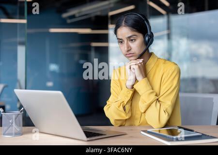 A serious young Indian woman in a headset is sitting at a desk in the office and looking thoughtfully at the laptop screen. Stock Photo
