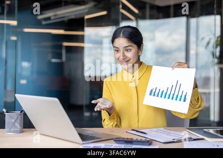 A young Indian businesswoman sits at a desk in the office and shows documents with graphs on a laptop over a video call. Stock Photo