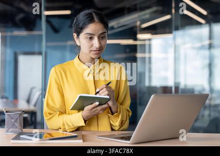 A young Indian woman sits at a desk in the office, looks at the laptop screen and makes notes in a notebook. Stock Photo