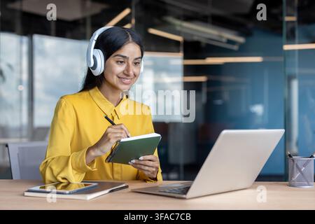 A young Indian woman wearing headphones sits at a desk in an office, working and studying on a laptop, looking at the screen and taking notes in a not Stock Photo