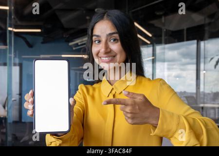 Close-up portrait of a young Indian woman in the office, holding a phone in her hand and pointing with her finger at a white mock-up screen. Stock Photo