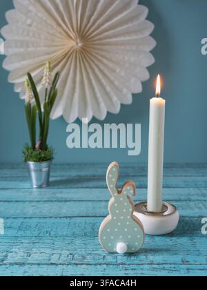Minimal Easter still life featuring a wooden bunny decorative candle small potted spring plant and large white paper flower on a blue wooden backgroun Stock Photo