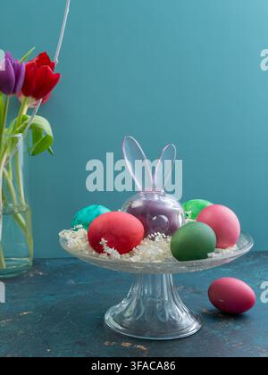 Elegant Easter still life featuring colorful decorative eggs arranged in a glass pedestal bowl with a wooden bunny and fresh tulips against a blue bac Stock Photo