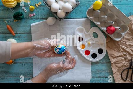 Creative Easter craft scene showing hands painting colorful eggs with brushes and a watercolor palette surrounded by additional eggs and craft supplie Stock Photo