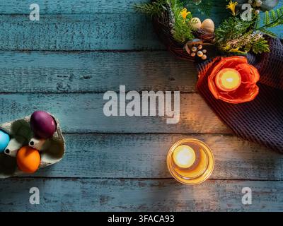 Festive Easter still life featuring a glowing candle decorative eggs spring greenery and a bright orange flower arranged on a blue wooden background. Stock Photo