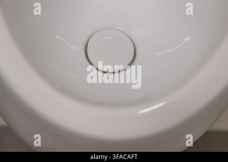 A detailed perspective of the smooth, reflective surface of a modern white ceramic urinal, highlighting the central drain mechanism Stock Photo