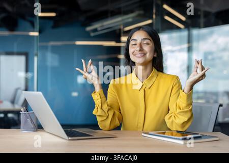 Relaxed young Indian businesswoman sitting at her desk in the office in lotus position, resting and meditating. Stock Photo
