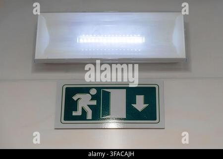 A bright LED emergency light fixture is mounted above a green exit sign indicating a path to safety Stock Photo