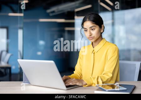 Serious young Indian businesswoman sitting at a desk in the office and working intently on a laptop. Stock Photo