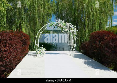 Luxury wedding venue featuring romantic floral arch on white wooden pier next to scenic lake. Elegant marriage ceremony location with exquisite white Stock Photo
