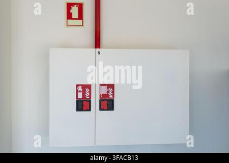 A white cabinet mounted on a wall, displaying symbols for a fire extinguisher and a hose reel, with a red pipe overhead Stock Photo