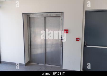 A contemporary stainless steel elevator entrance is shown next to a dark gray door with red emergency buttons mounted on the wall Stock Photo