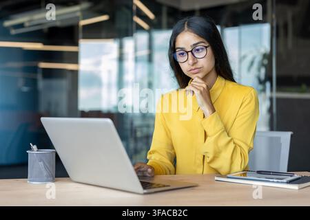 Serious young Indian businesswoman working in the office, sitting at the desk, looking thoughtfully and concentratedly at the laptop screen. Stock Photo
