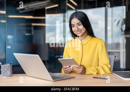 Smiling young Indian student studying at a laptop, holding a tablet in her hand and looking at the screen. Stock Photo