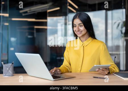 Smiling young Indian woman sitting at office desk, holding tablet in hand and using laptop. Stock Photo
