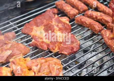 Close-up of marinated raw pork steaks, spiced chicken and minced meat sausages slowly roasting over hot burning charcoal embers on a shiny metal barbe Stock Photo