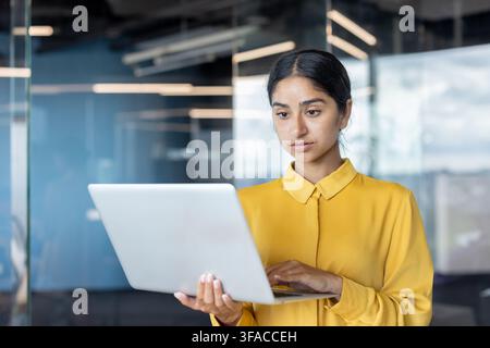Young Indian businesswoman standing in office, holding laptop and typing on keyboard. Stock Photo