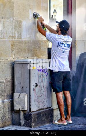 Young man with no face mask for dust protection using an electric power tool sander to clean an old stone wall - Tours, Indre-et-Loire (37), France. Stock Photo
