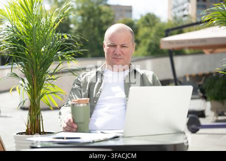 Mature caucasian man writing notes while using smartphone and laptop at an outdoor cafe, concept of business planning and remote work. Stock Photo