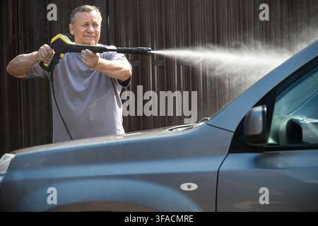 Mature Caucasian man using pressure washer to clean car outdoors, concept of home car wash, vehicle care, auto detailing and maintenance. Stock Photo