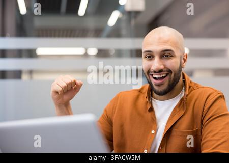 Happy professional showing excitement and joy, celebrating a victory or achievement while looking at a laptop computer in a modern office environment, feeling motivated and successful Stock Photo