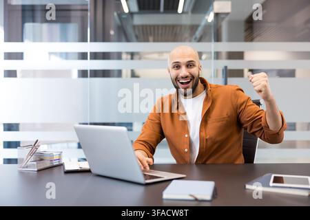 Young businessman celebrating success at his desk with laptop in a bright corporate office, smiling and pumping fist in triumph, confident and productive in modern workspace Stock Photo