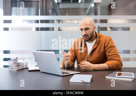 Bald man sitting at a desk, looking surprised and frustrated at his laptop screen while pointing, experiencing shock and confusion with technology issues in a modern workplace Stock Photo