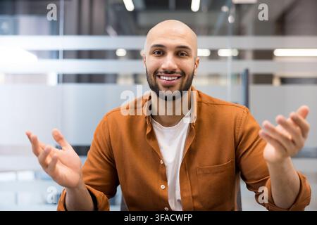 Young bald man with a beard smiling and actively gesturing with his hands during a virtual meeting, communicating remotely from a modern office workspace Stock Photo