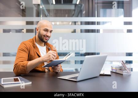Young man smiling while making notes in a notebook, actively engaged in planning and organizing tasks at a desk with a laptop in a contemporary office setting Stock Photo