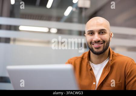 Young bald man with a beard smiling confidently while using a laptop, representing concepts of success, professionalism, productivity, and modern business in a comfortable office environment Stock Photo