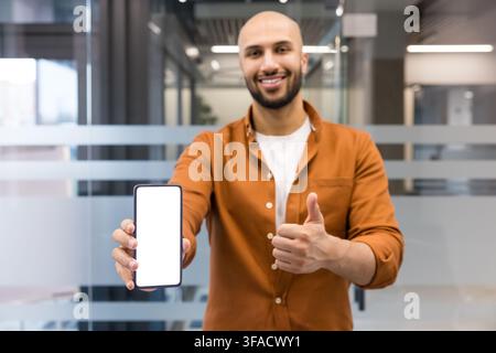 Happy young man holding up a mobile phone with an empty white display for mockup advertising, showing approval and satisfaction with a thumbs up gesture Stock Photo