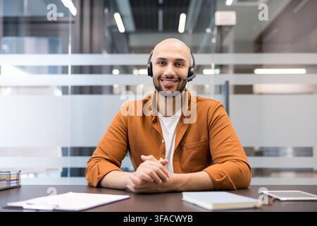 Smiling man wearing a headset and an orange shirt working at a desk in a modern office, providing telemarketing assistance and support to clients through online communication and virtual meetings Stock Photo