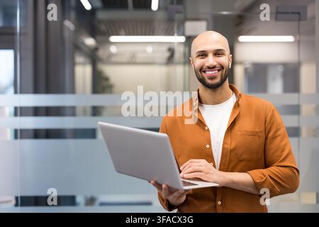 Confident young male developer smiling while holding a portable computer, standing in a contemporary office setting with glass partitions, representing innovation and career success Stock Photo