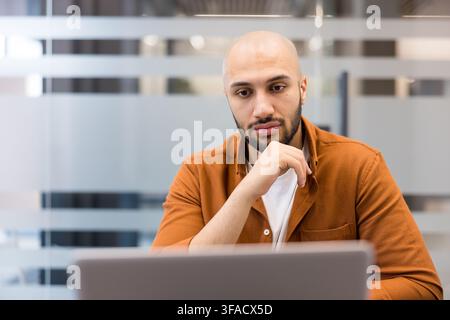 Young bald man looking at laptop screen, thinking deeply and concentrating on his work in a modern office, developing new strategies while focusing on problem-solving Stock Photo